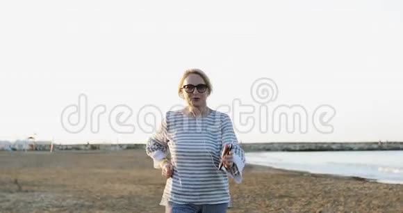 金发的老年妇女戴着太阳镜在海边沙滩上奔跑和跳舞视频的预览图