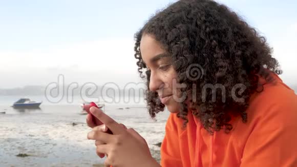 一位非洲裔美国少女身穿橙色连帽衫坐在海边用她的智能手机进行社交治疗视频的预览图