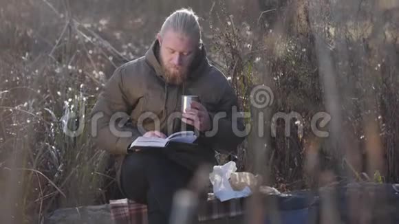 体贴的白种人坐在树干上看书喝着钢杯里的热茶孤独男游客视频的预览图