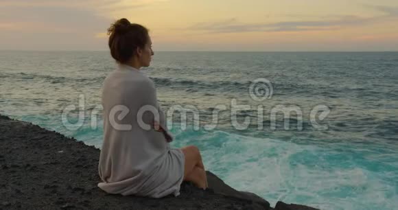 一个穿着格子布的女孩坐在码头上看着日落时的海浪视频的预览图