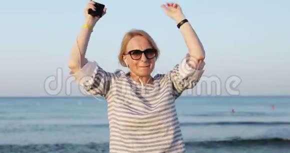 戴墨镜的金发老妇人正在海边散步和滑稽的舞蹈视频的预览图