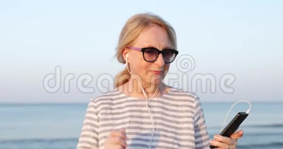 戴墨镜的金发老妇人在海边散步和跳舞在手机上听音乐视频的预览图
