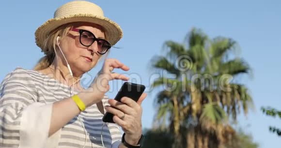 戴着太阳镜和草帽的金发老妇人在手机上选择了一首歌视频的预览图