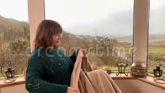 女人坐在一张铺着格子的舒适的柳条椅子上坐在山上一个灯光封闭的阳台上喝着热茶看着窗外视频的预览图