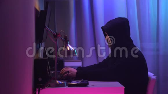 男性黑客坐在他的办公桌前闯入系统穿着黑色连帽衫的人把脸藏起来用霓虹灯做笔记本视频的预览图