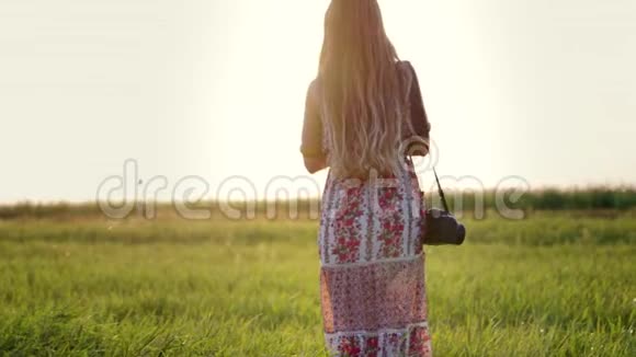 美丽的年轻嬉皮士女人肩上戴着相机在日落时分穿过田野视频的预览图