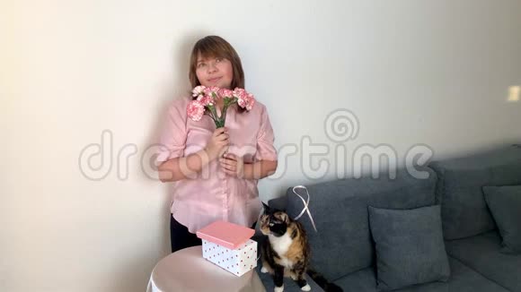 女人嗅着一束粉红色的康乃馨微笑着盒子里的礼物躺在圆桌上一只黑猫坐在灰色的沙发上视频的预览图