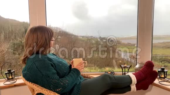 女人坐在阳台上舒适的柳条椅子上喝着热茶看着窗外享受和放松的概念视频的预览图