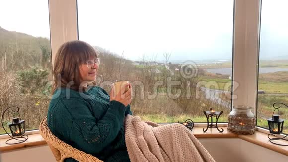 女人坐在阳台窗边舒适的柳条椅子上喝着热茶躲在毯子里看着窗外一个视频的预览图