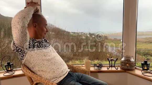男人坐在阳台上的宽窗旁舒适的柳条椅子上伸展休息和放松的概念视频的预览图
