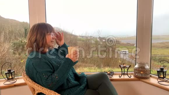 女人坐在阳台上舒适的柳条椅子上喝着热茶看着窗外这是一种享受和放松的概念视频的预览图