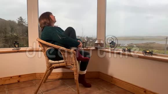 女人坐在一张舒适的柳条椅上坐在山上一个灯光封闭的阳台上享受着放松看着窗外紧紧抓住她视频的预览图