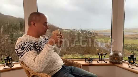 男人坐在阳台上舒适的柳条椅子上喝着热茶望着窗外享受和享受的概念视频的预览图