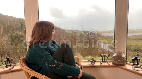 女人坐在一张舒适的柳条椅上坐在山上一个灯光紧闭的阳台上微笑着双腿穿着温暖的高袜子视频的预览图