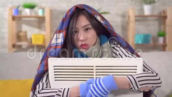 一个冻僵的年轻亚洲女人坐在客厅里的加热器的肖像视频的预览图