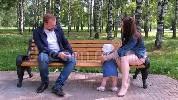 小女孩试图从妈妈到爸爸坐在公园的长凳上迈出第一步视频的预览图