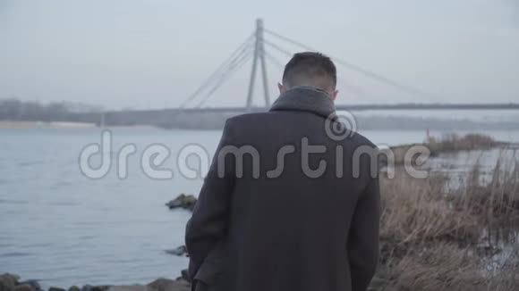 一个孤独的白种人走在岩石河岸边思考的中间镜头穿着优雅外套的年轻人的后景视频的预览图
