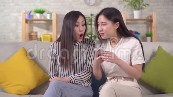 两个快乐的亚洲女孩坐在客厅的沙发上高兴地看着他们的智能手机视频的预览图