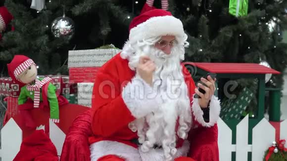圣诞老人在圣诞树附近的购物中心用他的智能手机工作视频的预览图