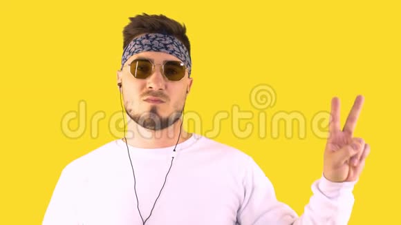 时髦的年轻人戴着时髦的眼镜和手帕在耳机上听音乐并显示手胜利标志黄色视频的预览图