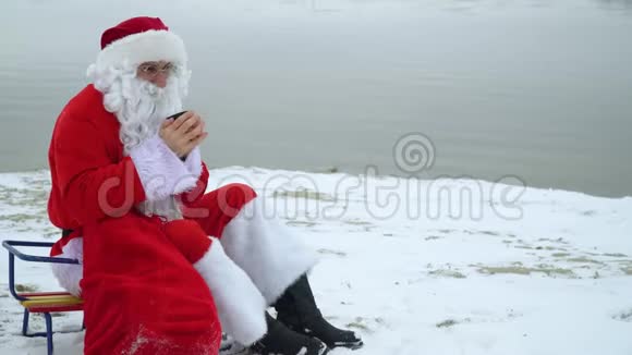 圣诞老人坐在湖的雪岸上拿着一袋礼物喝着热水瓶里的咖啡视频的预览图