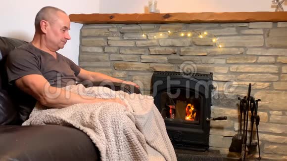 一位中年男子坐在一块烧着的石头旁边的皮革沙发上享受着温暖情感上的微笑和舒适的家庭概念视频的预览图
