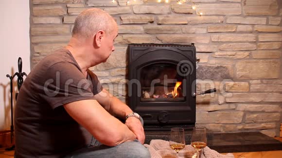 一个男人坐在燃烧的壁炉前的地板上脚边是两杯酒一个浪漫的夜晚的概念视频的预览图