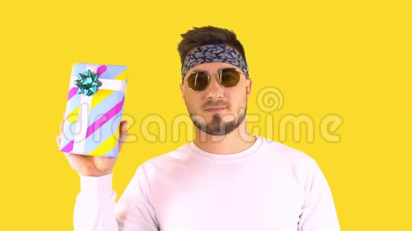 时尚的年轻人在时髦的眼镜班达纳显示拇指在盒子与礼物后面是黄色背景节日气氛视频的预览图