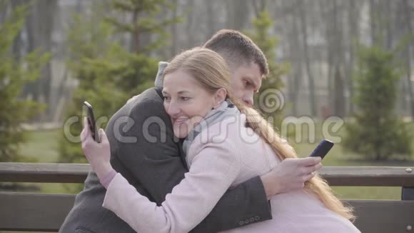 使用智能手机拥抱高加索成年夫妇的侧面视图金发女人和黑发男人利用社交媒体约会视频的预览图