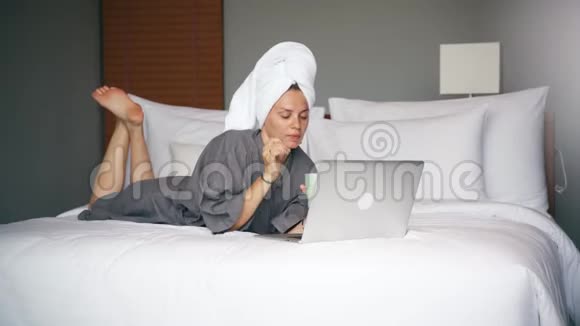 年轻的白种人女士在酒店房间里坐在床上时使用笔记本电脑视频的预览图