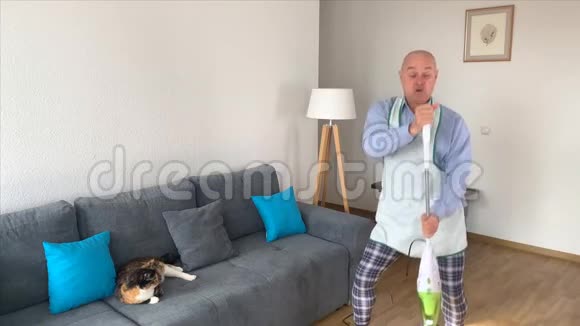 55岁的秃头男穿着家居服在沙发附近的房间里用围裙吸尘地板家庭分配的概念视频的预览图