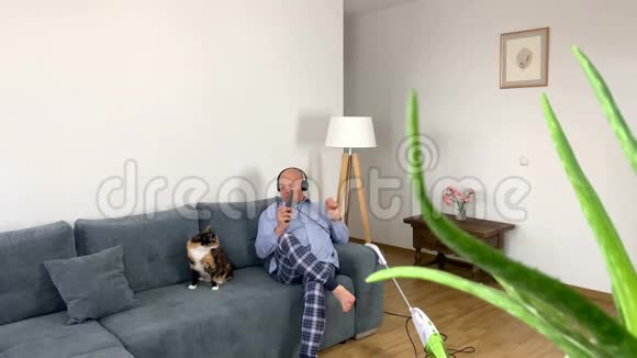 55岁的秃头男人穿着家居服围裙坐在沙发上靠近一只猫和一个吸尘器家庭琐事的概念视频的预览图