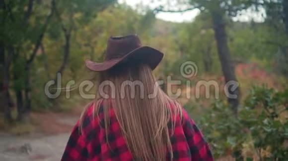 一个长发女孩穿着一件黑色和红色的衬衫戴着一顶牛仔帽正穿过树林视频的预览图