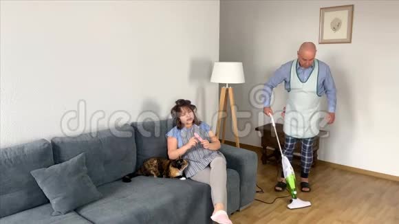 55岁的秃头男人穿着家居服在沙发附近的房间里用围裙吸尘地板女人男人坐在沙发上而不是视频的预览图