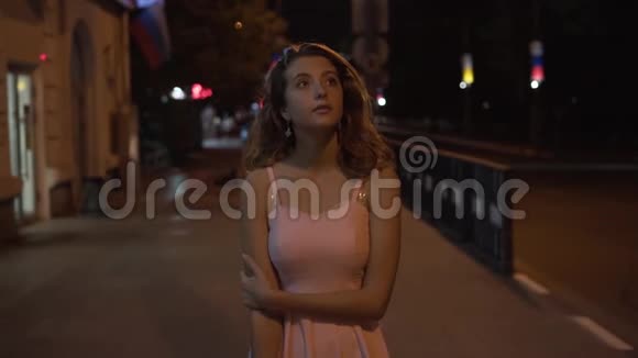 一个孤独的女孩穿着一件粉红色的连衣裙卷发在晚上沿着街道走视频的预览图