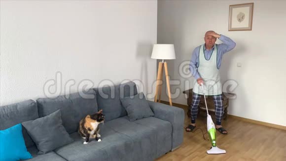 55岁的秃头男穿着家居服在沙发附近的房间里用围裙吸尘地板家庭分配的概念视频的预览图
