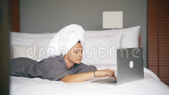 年轻的白种人女士在酒店房间里坐在床上时使用笔记本电脑视频的预览图