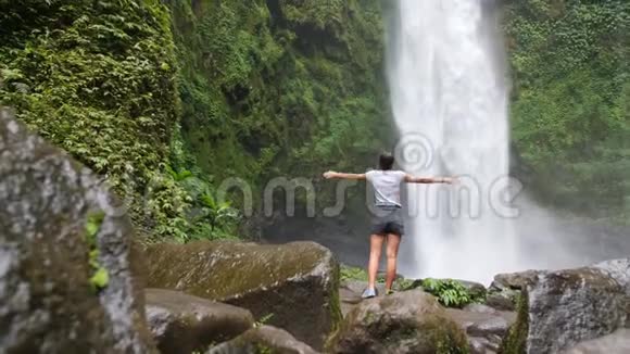年轻的旅行者女孩拿起武器在惊人的丛林瀑布在巴厘岛印度尼西亚4K慢镜头电影旅行视频的预览图