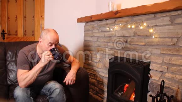 一个喝着咖啡和茶的中年男人坐在一块烧着的石头旁边的皮革沙发上享受着温暖有感情地微笑着视频的预览图
