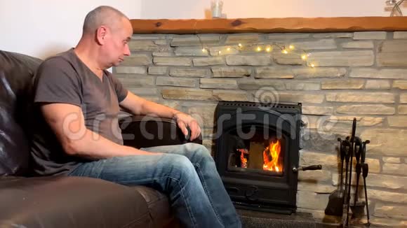 中年男子坐在炽热燃烧的壁炉旁双臂伸到炉火旁冬天的心情概念视频的预览图