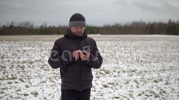一个穿着破旧冬装的男人冬天站在一个乡村的田野里进行在线视频聊天视频的预览图