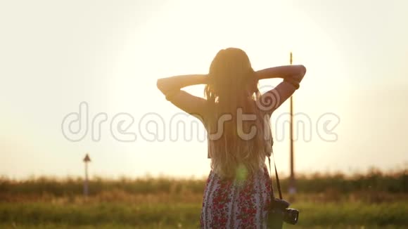 一个棕色头发的女人的肖像她穿着一件连衣裙带着相机在夕阳下一片高草的田野里视频的预览图