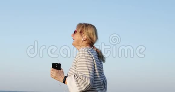 六十岁的快乐女人戴着耳机做早操视频的预览图