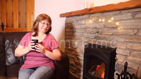 一个女人端着一杯咖啡喝着茶坐在一块烧着的石头旁边的皮革沙发上享受着温暖情感上的微笑享受着这个概念视频的预览图