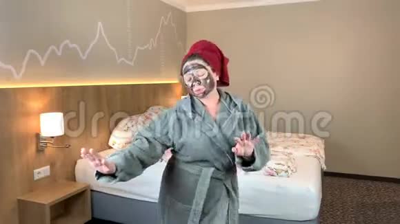 戴着黑色化妆面具的女人头上戴着毛巾穿着浴袍在床边跳舞有面部护理的概念视频的预览图