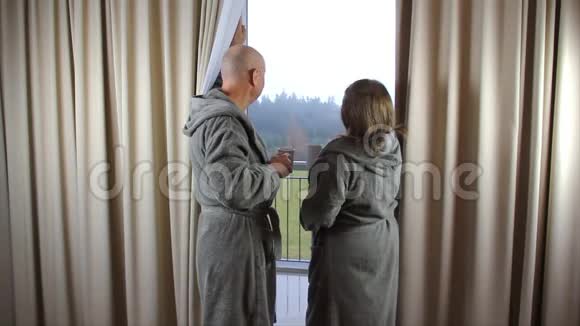 积极的一对夫妇穿着浴袍的男人和女人在酒店房间的窗户边喝咖啡度假的概念旅行视频的预览图