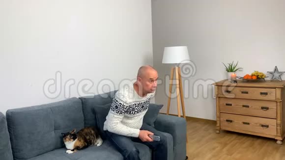 穿着北欧白色毛衣的成年男子坐在猫旁边的沙发上看电视视频的预览图