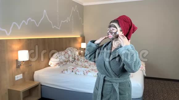 戴着黑色化妆面具的女人头上戴着毛巾穿着浴袍站在床边有面部护理的概念视频的预览图