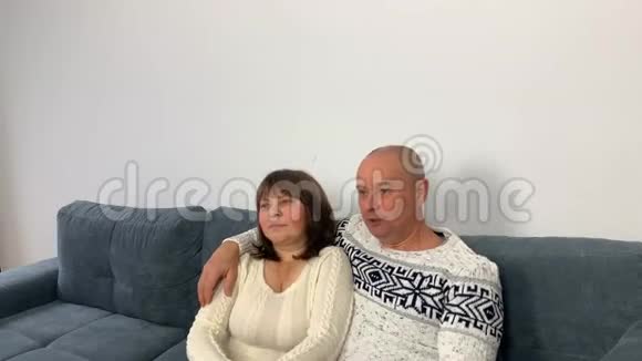 穿着斯堪的纳维亚白色毛衣的成年男子和坐在沙发上看电视节目的妇女视频的预览图