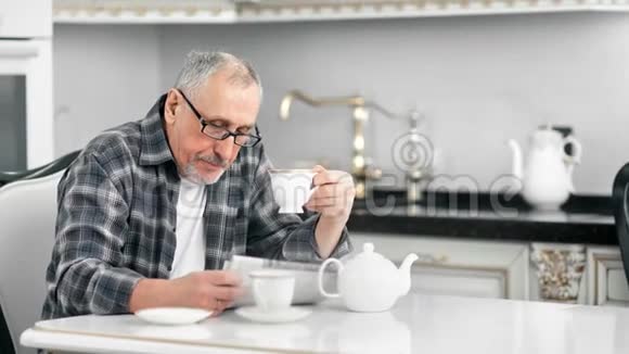 自信的中年留胡子的男人在白色厨房里看报纸时喝茶视频的预览图